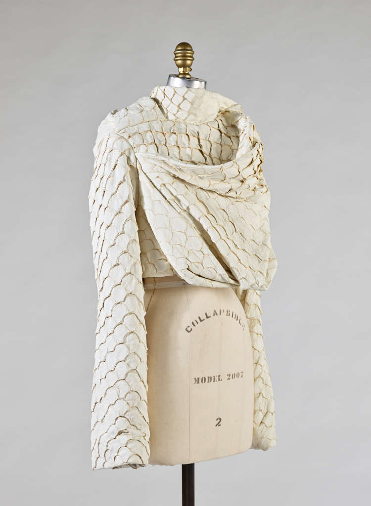 A cream colored, draped garment with wide sleeves and gold-accented scallop designs, fitted on a mannequin torso.