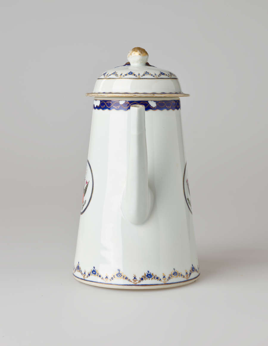 A white coffee pot with dark blue and gilded decorations, a straight spout, angular handle, and heraldry imagery in a circle on the body of the coffee pot.