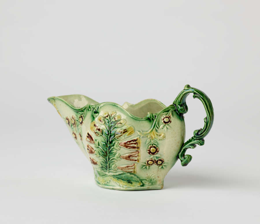  A short boat shaped pitcher green, cream, and brown in color with a sculptural handle and floral decorations.
