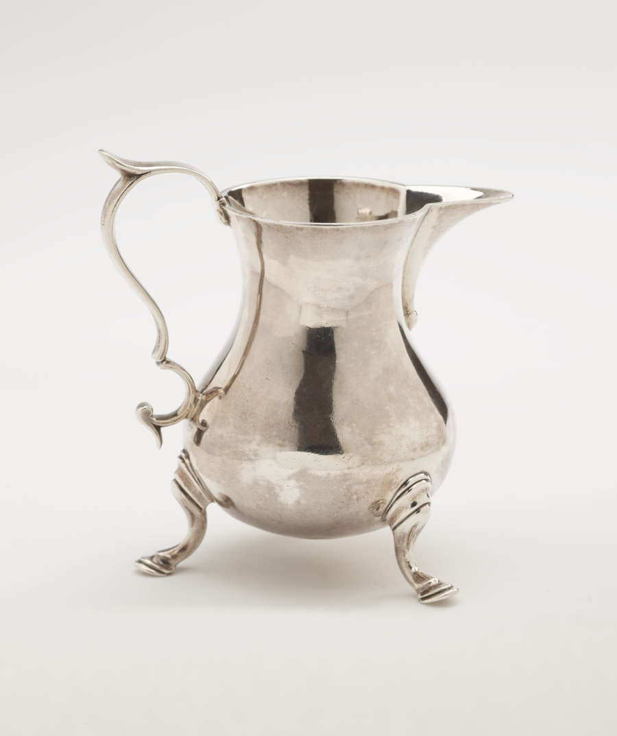 A silver creamer with a sculptural handle and three protruding feet.