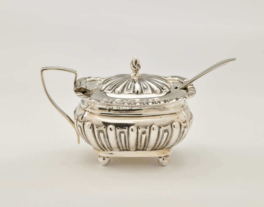 A silver, glass, and gilded mustard pot with a protruding spoon. The lid is hinged to the angular handle. The decorations are raised.