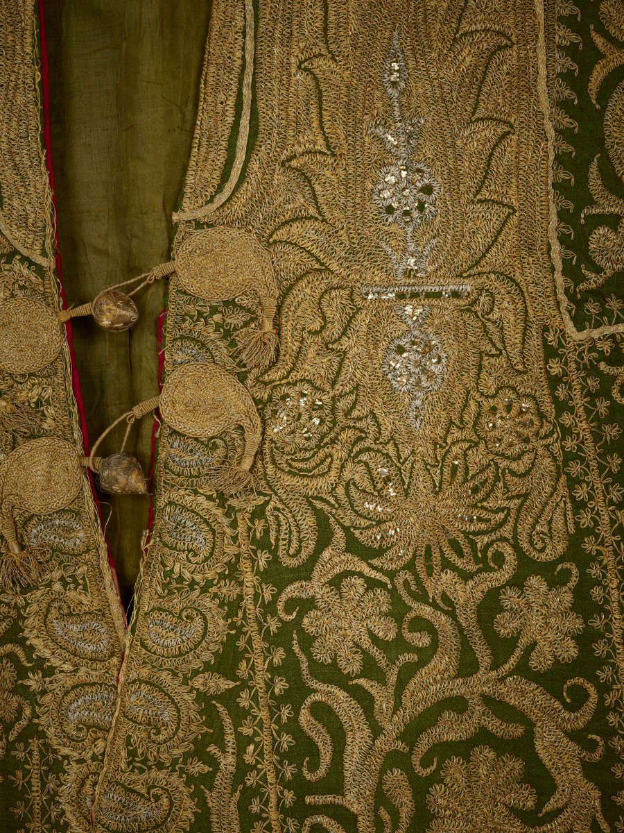 Close-up of a green coat’s neckline, highlighting the intricate gold and silver floral embroidery and paisley strip along the selvedge. It is secured with cloth bead and loop buttons.