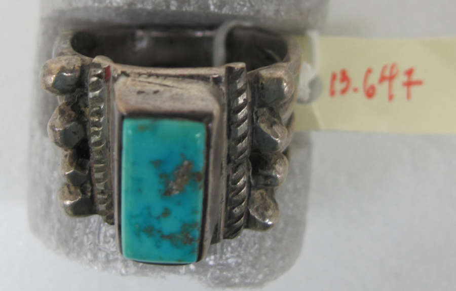 Image in storage of a silver ring with a rectangular turquoise stone-setting embellished rope-like and nature-inspired decorative elements. Next to the ring is a yellow tag labeled ‘13.647’.