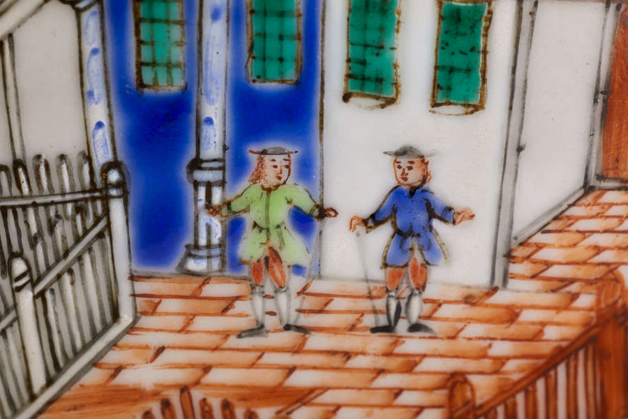 A close-up of a painted ceramic depicting two figures dressed in blue and green, standing on a brick pathway near a building with green windows and blue doors.