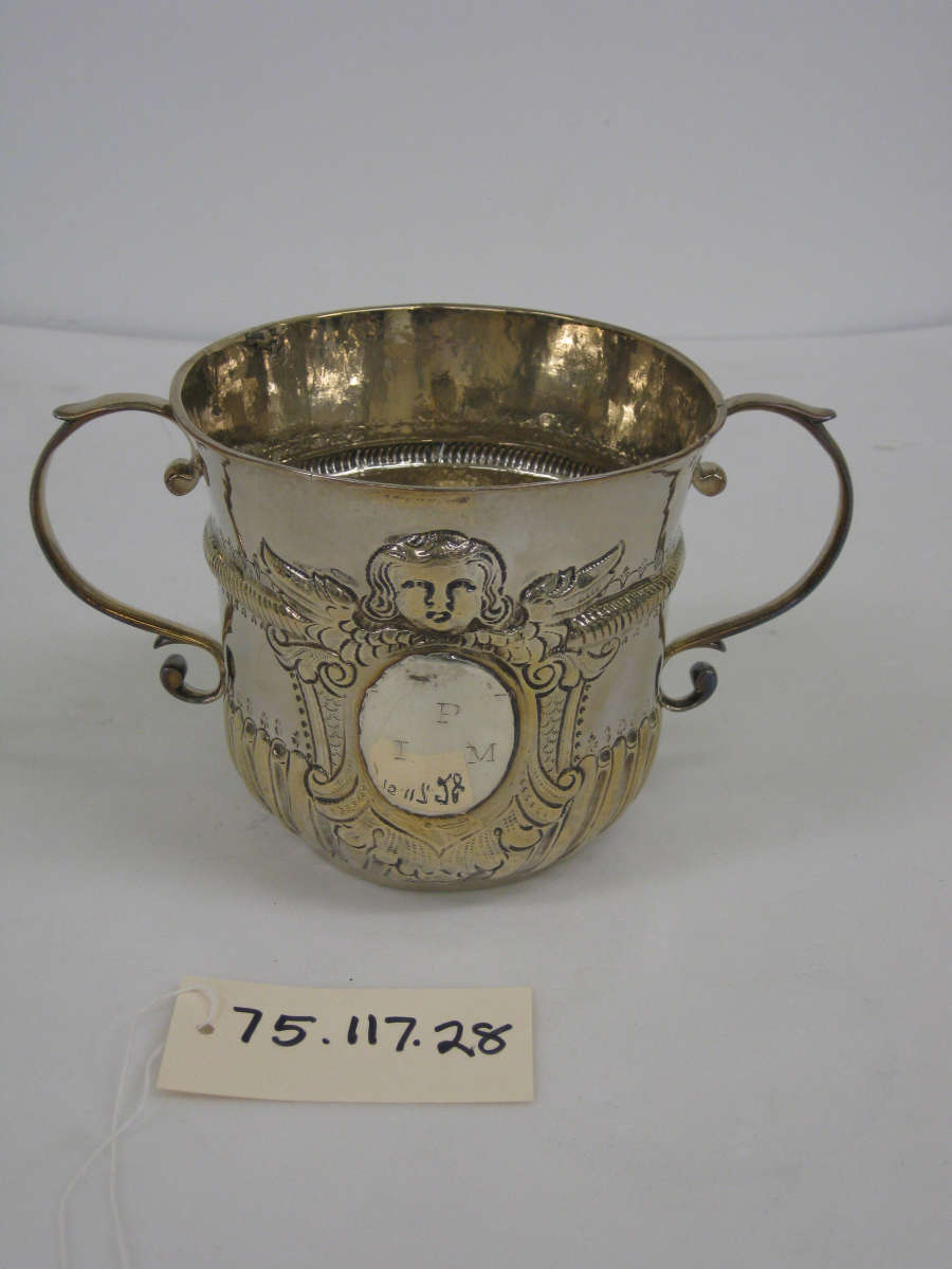 A silver cup with symmetrical handles, swirling raised designs and a small raised face on the upper half of the cup.
