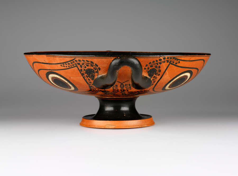 Side view of a terracotta and black cup with a wide mouth, short black neck, and orange base. Its body features circular motifs rendered in black and white. 