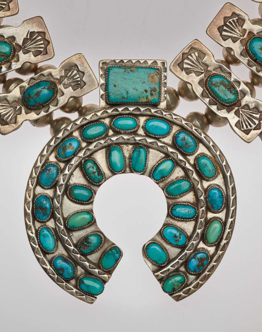 Detail-view of a horseshoe-shaped pendant of a turquoise and silver necklace. The turquoise stones are set in two concentric circles with decorative silver borders with a rectangular stone-setting atop it.