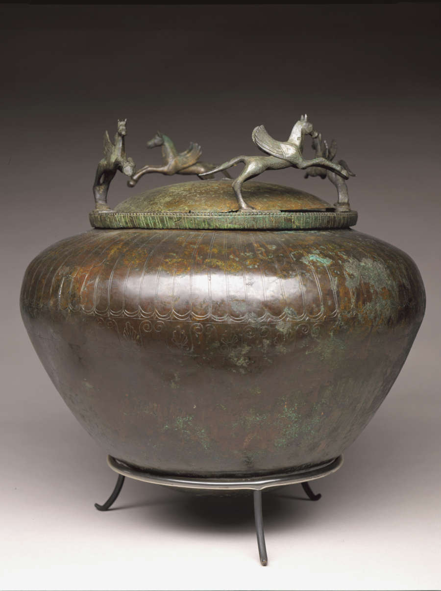 A brown-green patinated metal bowl with decorative engravings on its onion-shaped body, sitting on a three-legged stand. Metal-cast winged horses dance along the rim of the bowl’s domed lid.