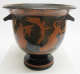 Stout black and terracotta bowl with two handles, decorated with floral motifs wrapped around the cup’s mouth as well as  illustrations of humans, cherubs, and satyrs gesturing towards one another.