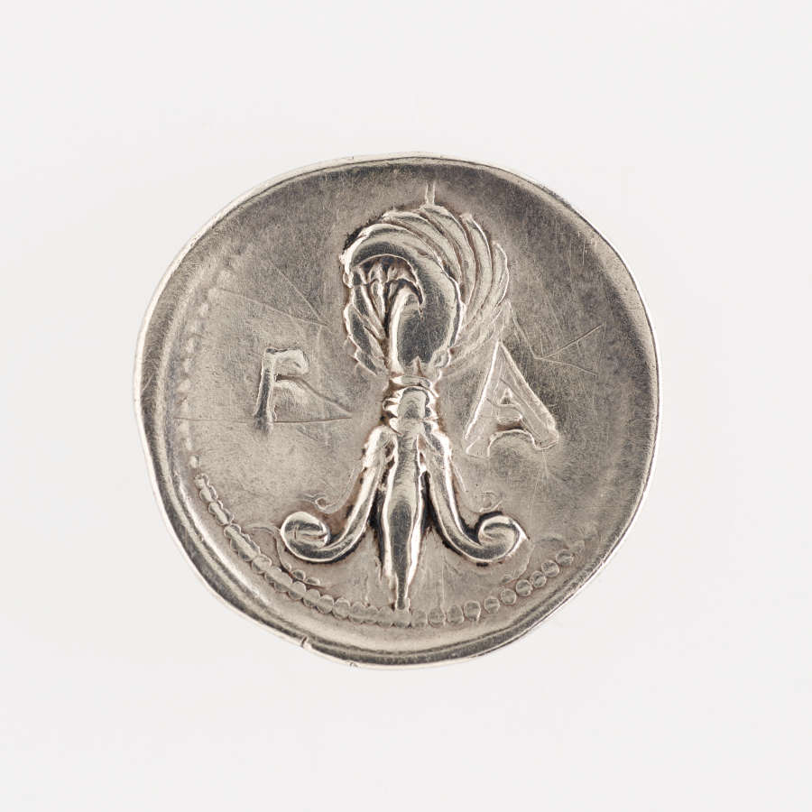 Irregular round silver coin embossed with the image of a feathered fleur-de-lis and the letters “F” and “A” on either side of it. It is framed by a dotted border.