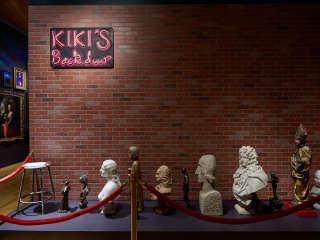 A brick wall bears a sign saying “Kiki’s Back Door.” Waiting on the ground behind a red velvet rope is a row of portrait busts and historical figurative artworks.
