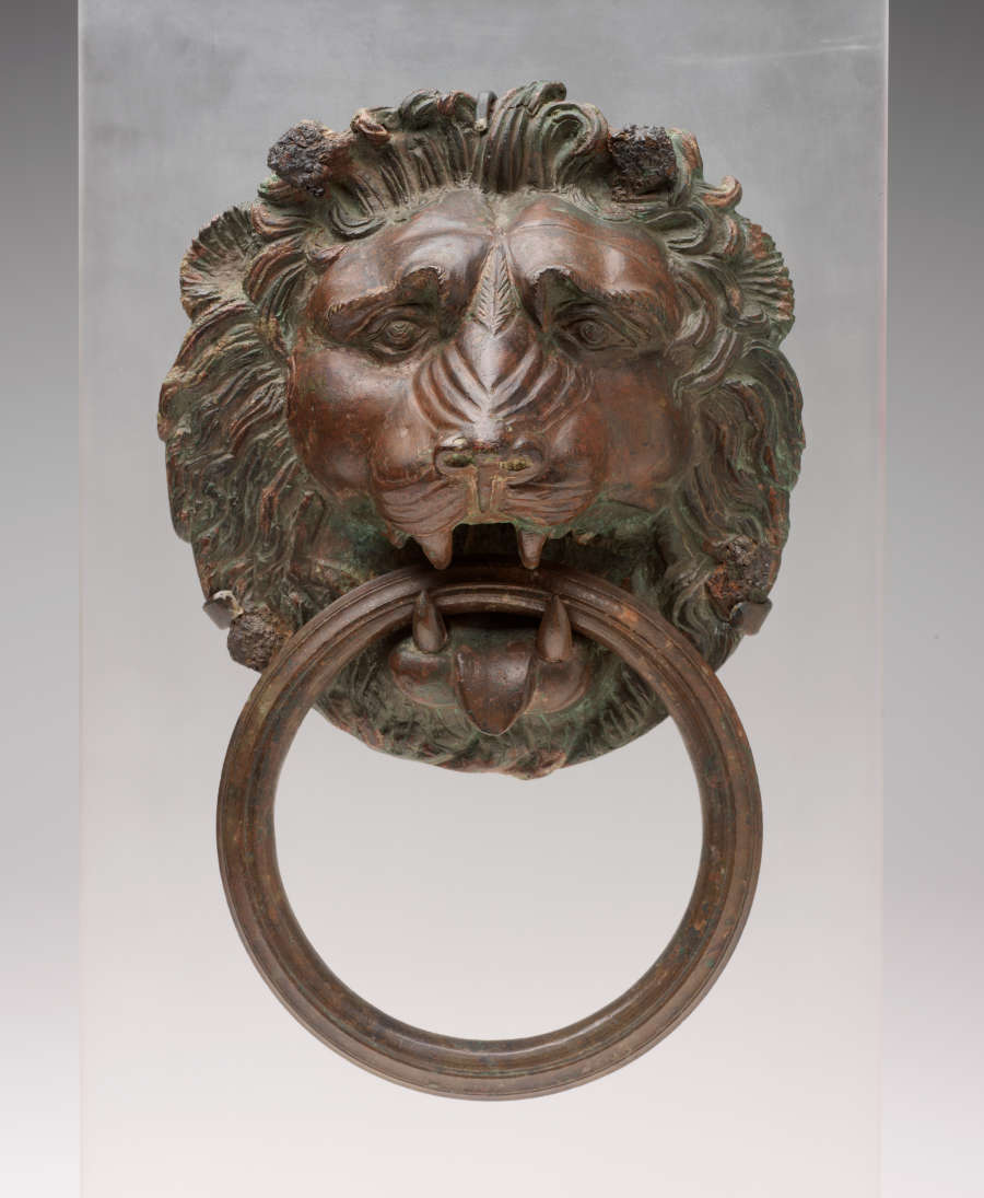 Gray-brown, slightly glossy sculpture of a lion’s head biting a thick, large, circular handle. The lion’s mane is well manicured and its face bears a quizzical brow.