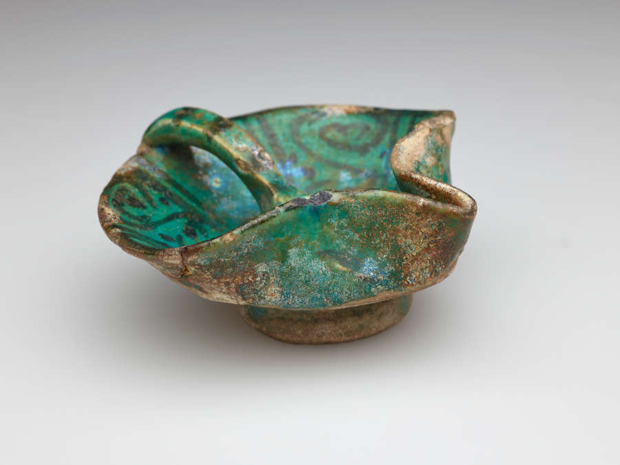 An irregularly shaped blue-green iridized vessel with dark designs. Rounded on one side with a handle that comes to the center of the vessel. The other side has a folded spout-like edge.