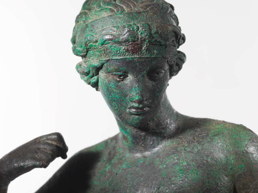 Detail view of a bronze statue of a nude woman at an angle. Visible are the details of her face, hair and clenched fist curled towards her shoulder.