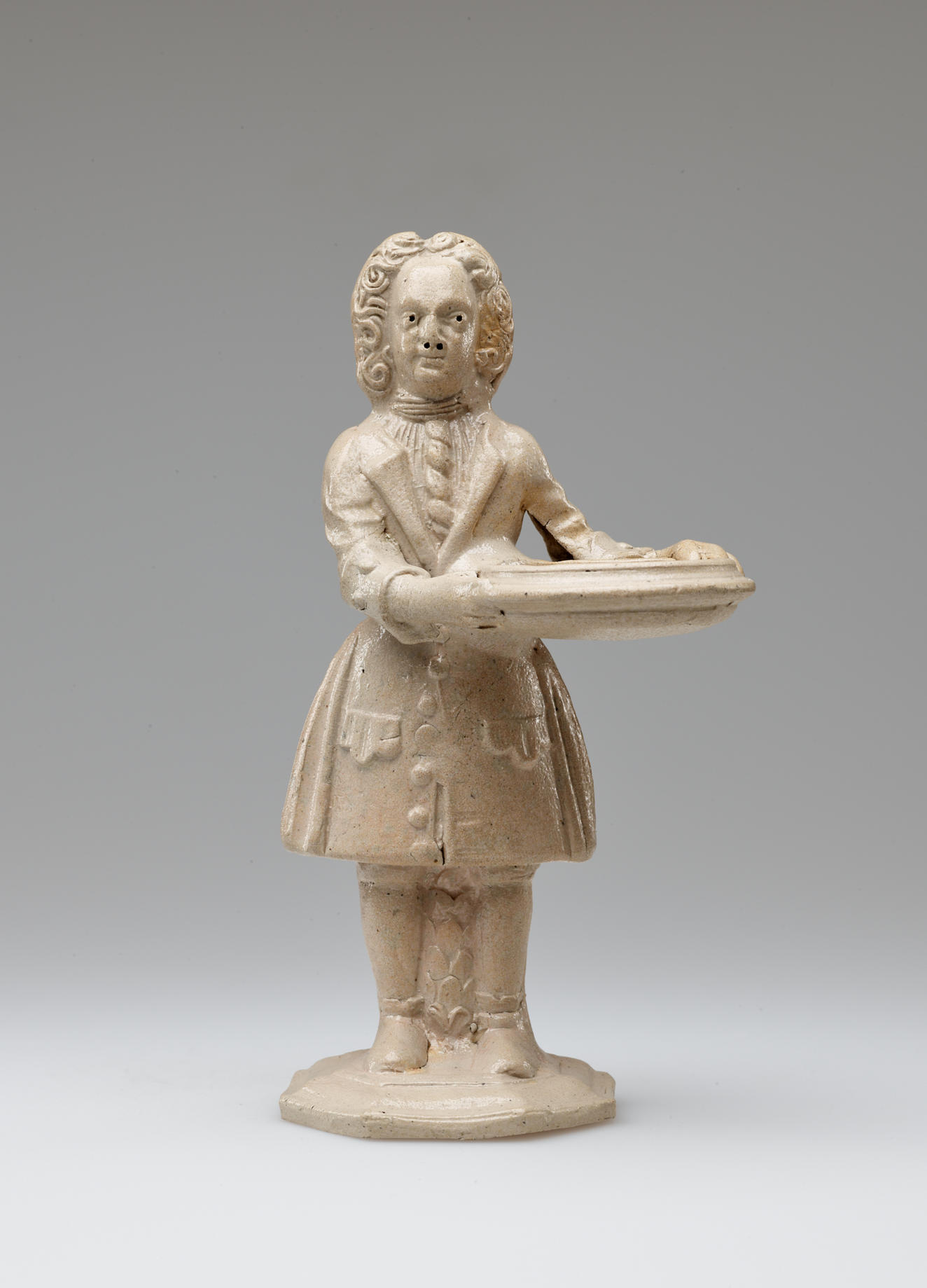 A cream colored sculptural figure holding a tray, dressed in historical clothing with tightly curled hair.