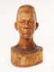 Front view of a glossy, carved wooden bust depicting the head of a male figure with afrocentric facial features. He has a stern expression and an intense gaze.