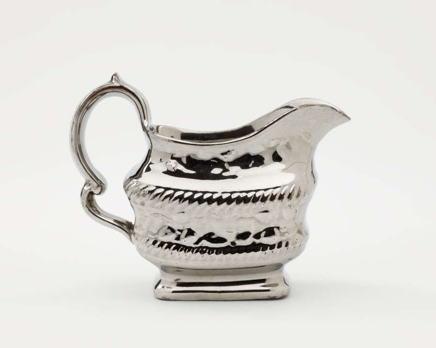  A silver creamer with a decorative handle and a rounded square body with sculptural decorations.
