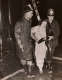 Black and white photo of a person in a white dress being assisted by two firefighters wearing long coats and helmets and holding a hose. A ladder is in the dark background.