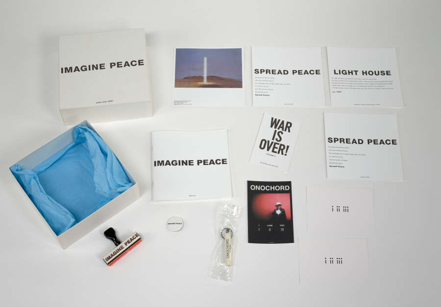 Photo of a box and various peace-themed items, including: a stamp, keychain, pin, and flyers that read “War is Over,” “Spread Peace,” “Light House,” and “Imagine Peace.” The box top reads “Imagine Peace” and box base has blue tissue paper inside. 