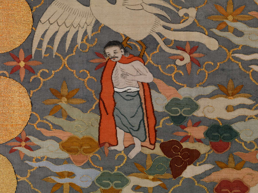 Robe’s back detail. Below a white bird and beside golden circles is a man looking downwards, against a dark background with a diagonal grid, within each square is a flower.