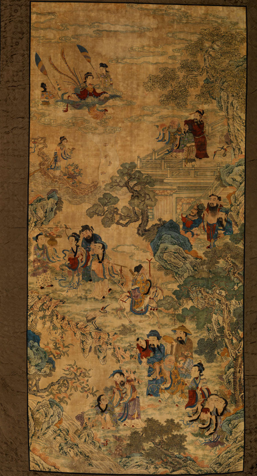 Vertically long rectangular scroll with a thick brown border. It features an illustration of several people engaging with another amongst trees, houses, and in boats on water.