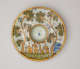 Round plate decorated with an image of buildings and trees. There is a concave well in the center of the form.