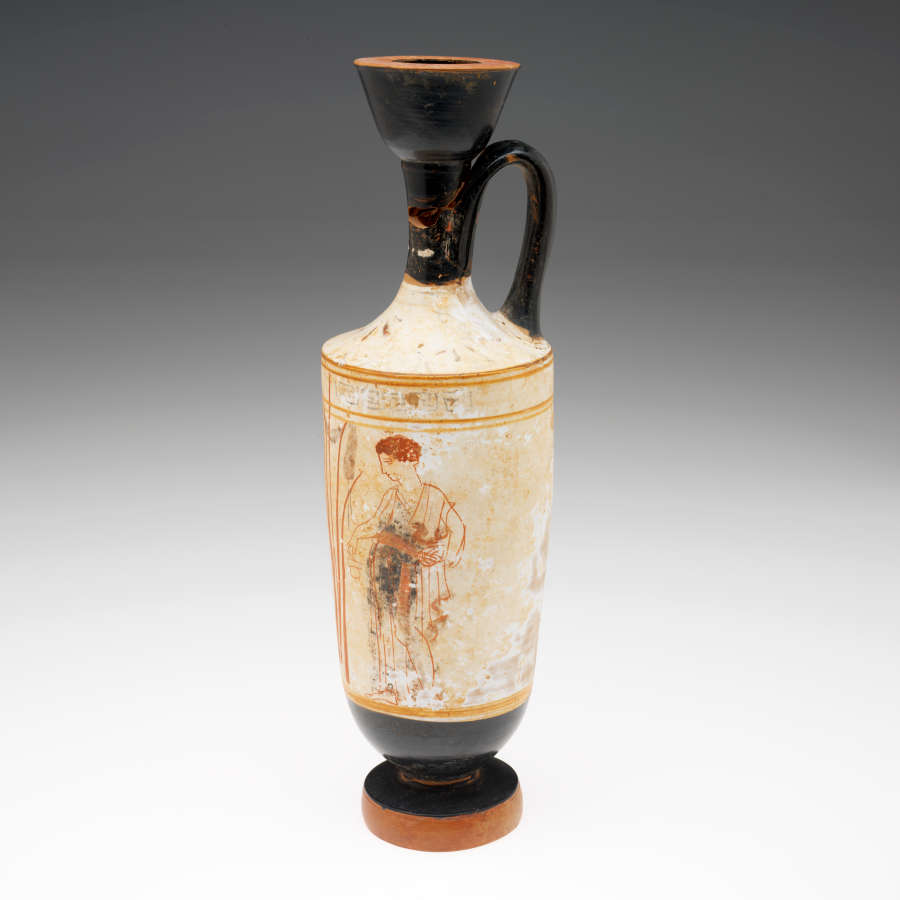 Tall terracotta vase, with a black fluted neck, wide mouth, thick base and handle. Its white body features fragmented illustrations rendered in red of a man facing reeds.