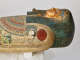 Side view of a coffin, featuring a gilded face with blue-lined eyes and a sculpted false beard. A painted falcon with a red sun is painted on the shoulder.