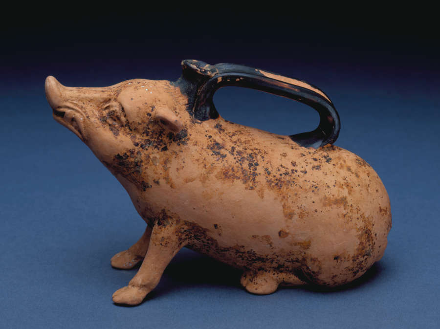 Side-view of a terracotta container shaped as a seated boar with an upturned snout looking upwards on a blue background. Along its back is a chipped long black handle. 