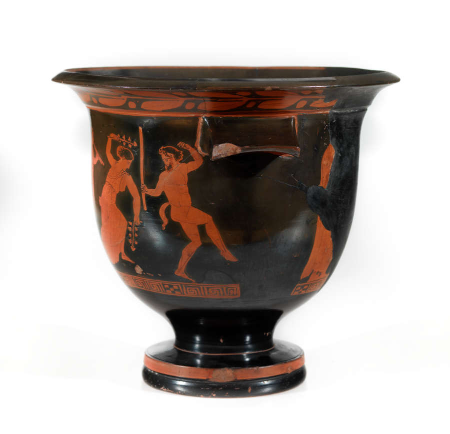 View of a stout black bowl with a wide striped foot and flared patterned mouth. Visible is one sloping handle, and the terracotta illustration of humans chasing satyrs.