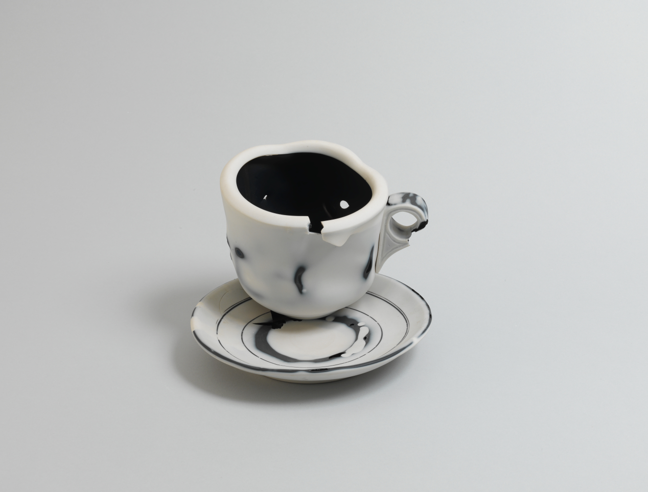 Marbled black-and-white ceramic teacup with an irregular, chipped rim and tiny circular handle, resting on the edge of a matching white saucer decorated with thin concentric black rings.