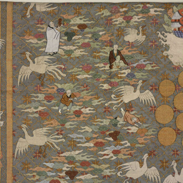 Embroidery depicting cranes in flight against multicolored clouds holding robed figures, and gold discs against a blue background with gold borders.
