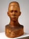 Front view of a carved wooden bust depicting the head of a male figure with afrocentric facial features. He has a stern expression and an intense gaze.