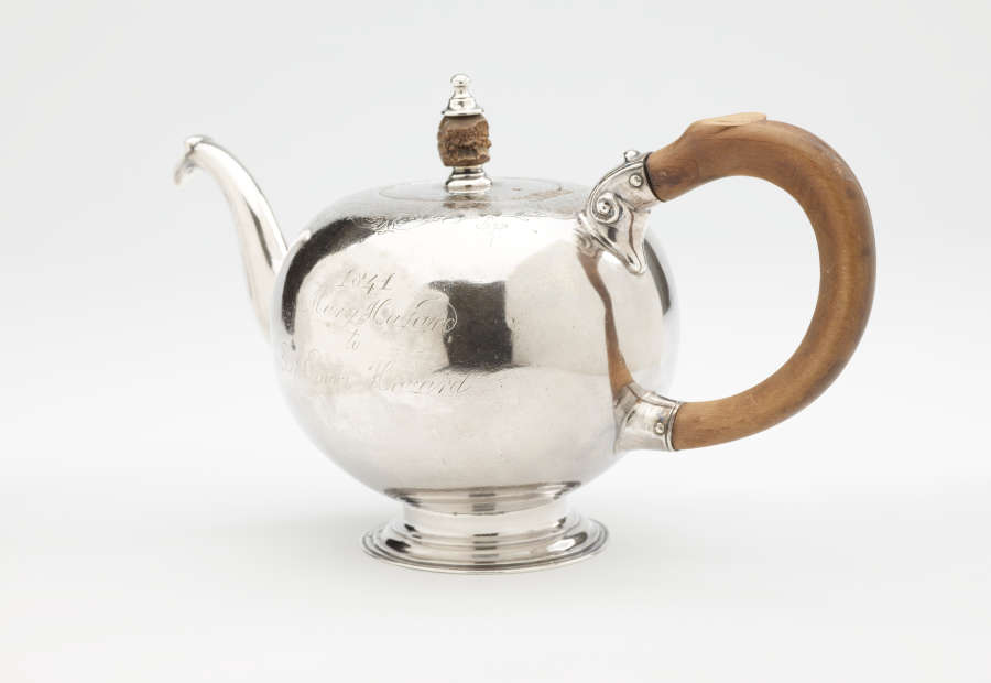 A bulbous silver teapot with a wooden finial and handle connected with decorative silver pieces. There are delicate engraved marks on the body and lid.