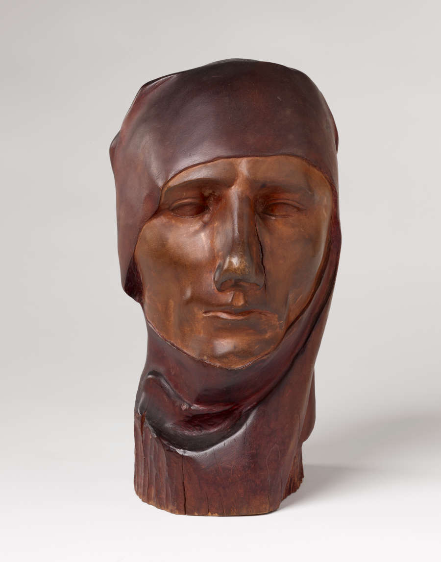 Front view of a vibrant, carved wooden sculpture depicting a serene face wearing a cloth draped around its head. The bust features smooth contours and a crack alongside its nose.