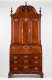 A tall mahogany cabinet and desk structure with a polished finish, slant-front design, intricate shell-shaped carvings, brass keyholes on all drawers, and a grand arched top decorated with ornamental details.