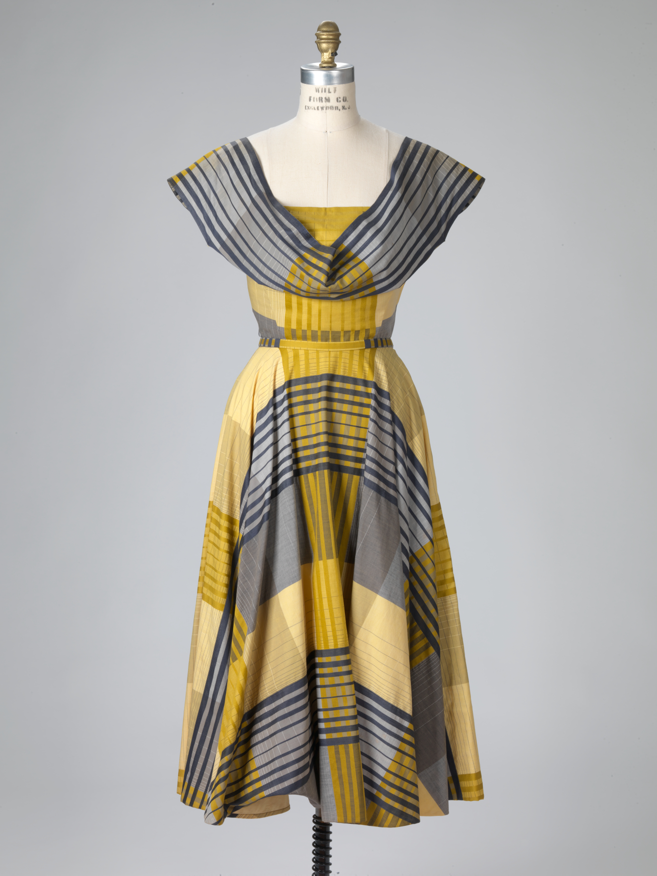 A knee-length mustard yellow and gray plaid dress with navy striped lines, pleated skirt, wide draped neckline merging into cap sleeves, and a cinched waist displayed on a mannequin.