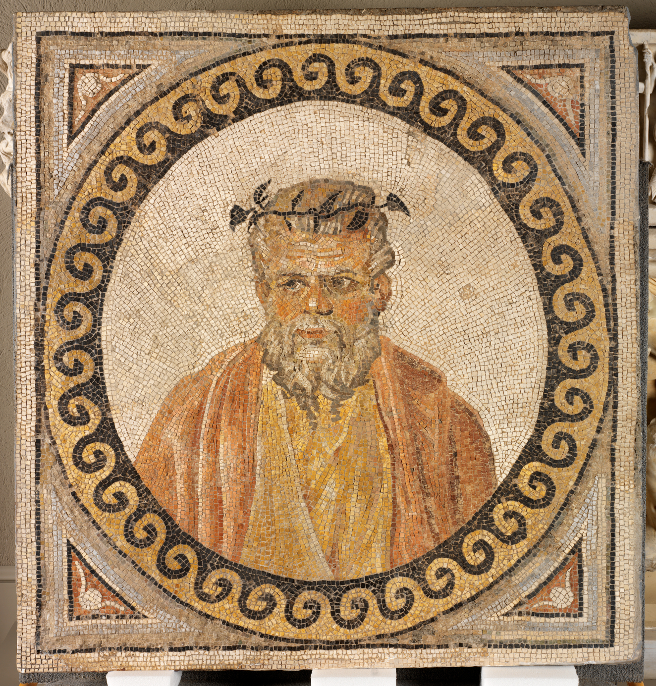 Mosaic portrait of a bearded man in a gold and red robe, framed by a circular black and yellow wave pattern.
