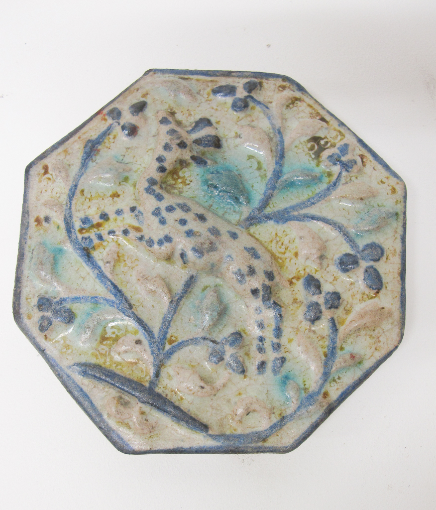 White octagonal tile with relief of intricate, flowing dark and light blue floral patterning overlaid with a stippled form of a gazelle at its center and bordered in blue.