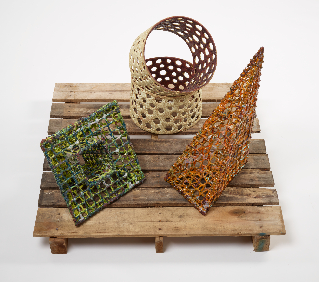 Three ceramic, netted, geometric forms, in green, orange and beige, creating cylinder, pyramid, and funnel forms on a wooden pallet.