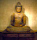 A wooden Buddha statue sitting cross-legged on a platform, hands resting in the lap, with a serene expression and closed eyes, warmly lit against a neutral backdrop.