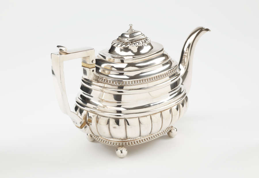 A silver teapot with a decorative angular handle, a rounded square body and spout with sculptural decorations.