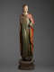 A tall, weathered wooden statue of a robed figure with a serene expression, draped in green and red garments, standing upon an octagonal base.