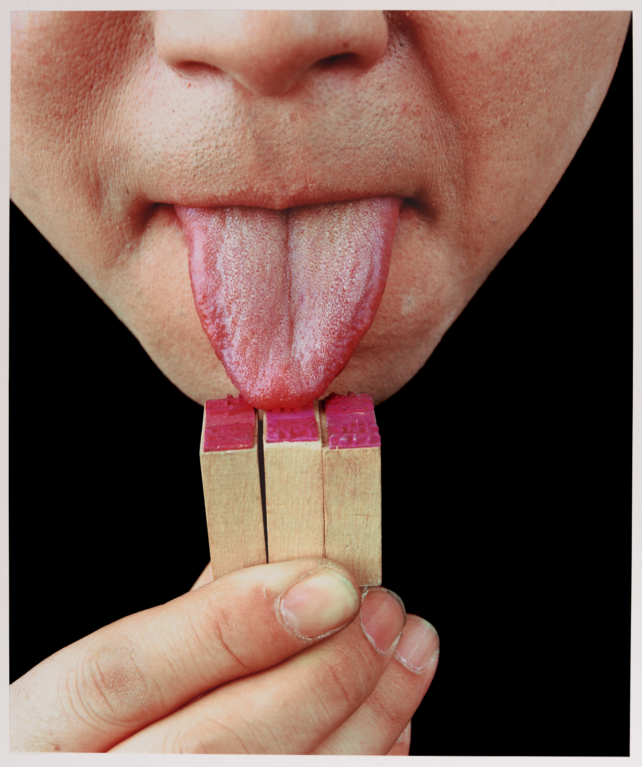 A close-up photograph of the bottom half of a huma face with a large tongue sticking out and touching three wooden stamp blocks, held up by the person's hand on a totally black background. 