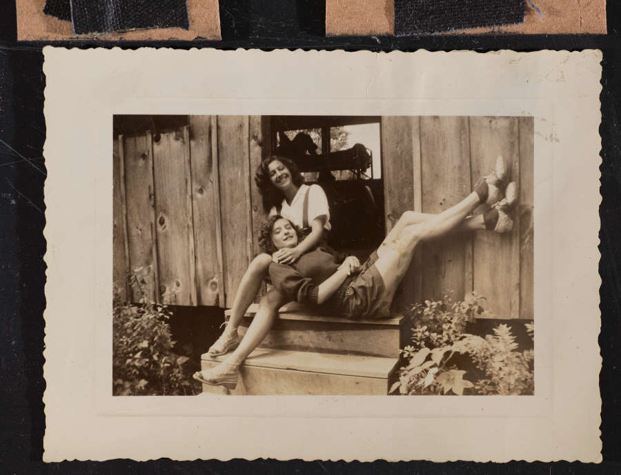 Vintage snapshot of two light-skinned young women mugging for the camera at the doorway of a cabin. One woman lies across the top step, her head in the other’s lap.