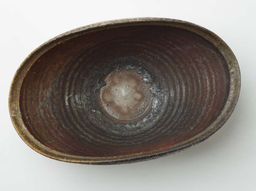 Top view of an oblong shaped stone bowl with a thick protruding rim. Its interior is dark orange blending into greens and whites near the center.