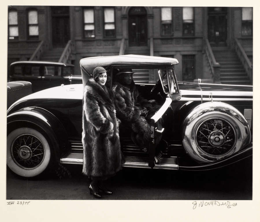 Black and white photo of a smiling woman in a hat and fur coat, beside an elegant car. A man inside the car also wears a hat and fur coat.