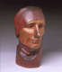 Three quarter front view of a vibrant, carved wood sculpture depicting a serene face, with closed eyes, wearing a draped cloth around its head. The bust is smooth and polished.