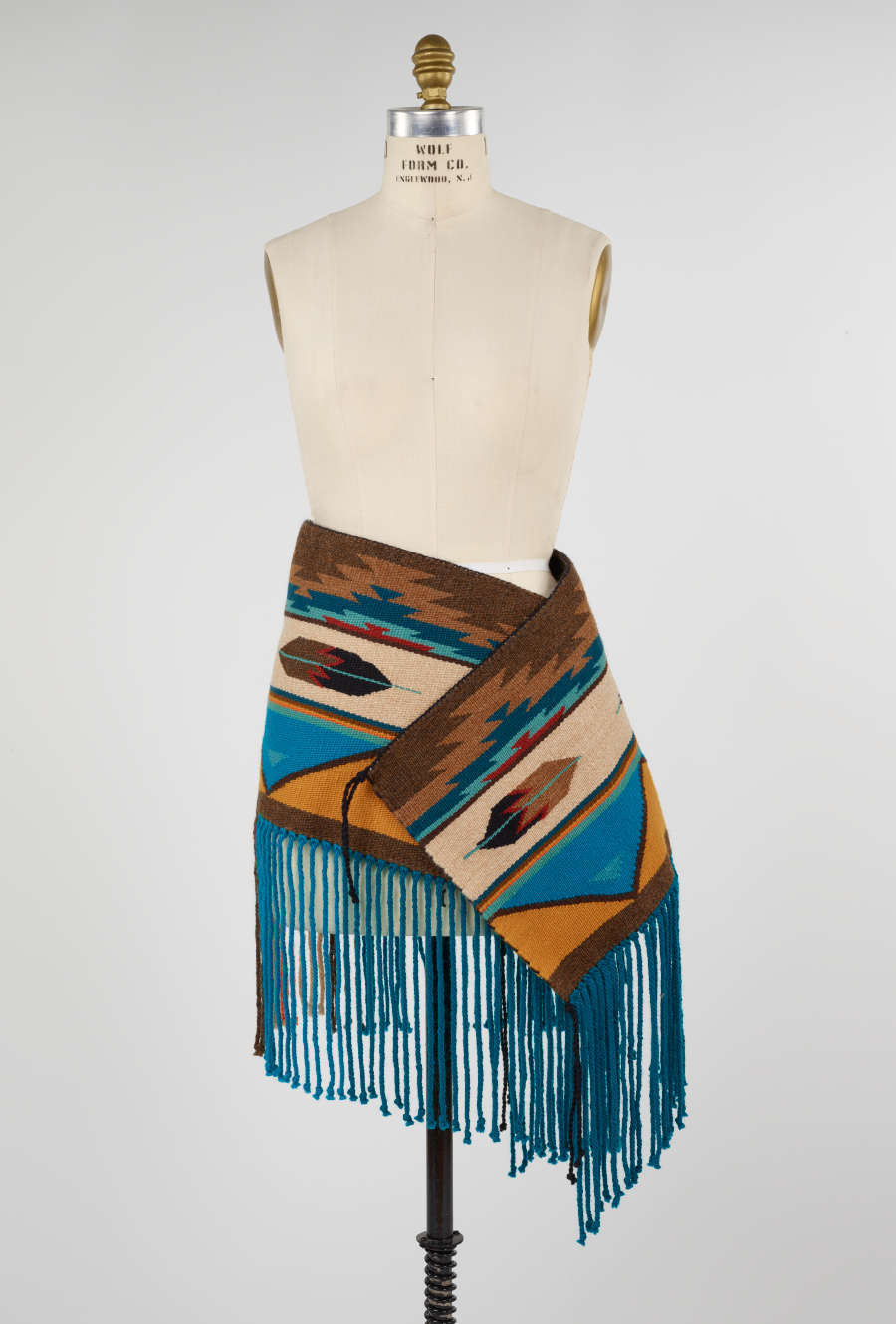 Turquoise, beige, and yellow geometric patterned woven fabric with fringe, wrapped around the hip of a mannequin. The turquoise fringed ends are the same length as the woven fabric. 