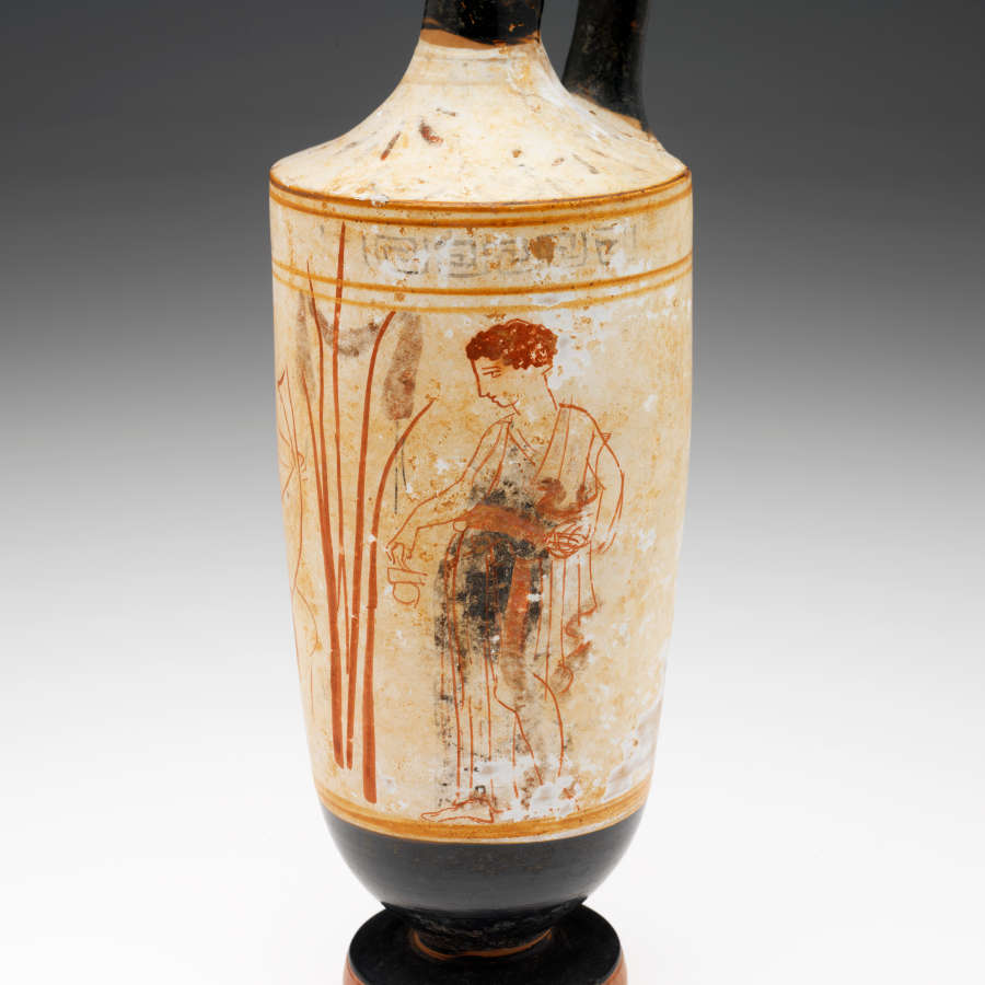 Close-up of the vase’s cream body. Visible are red illustrations of a man holding a small object standing besides a cluster of reeds, bordered by golden stripes.